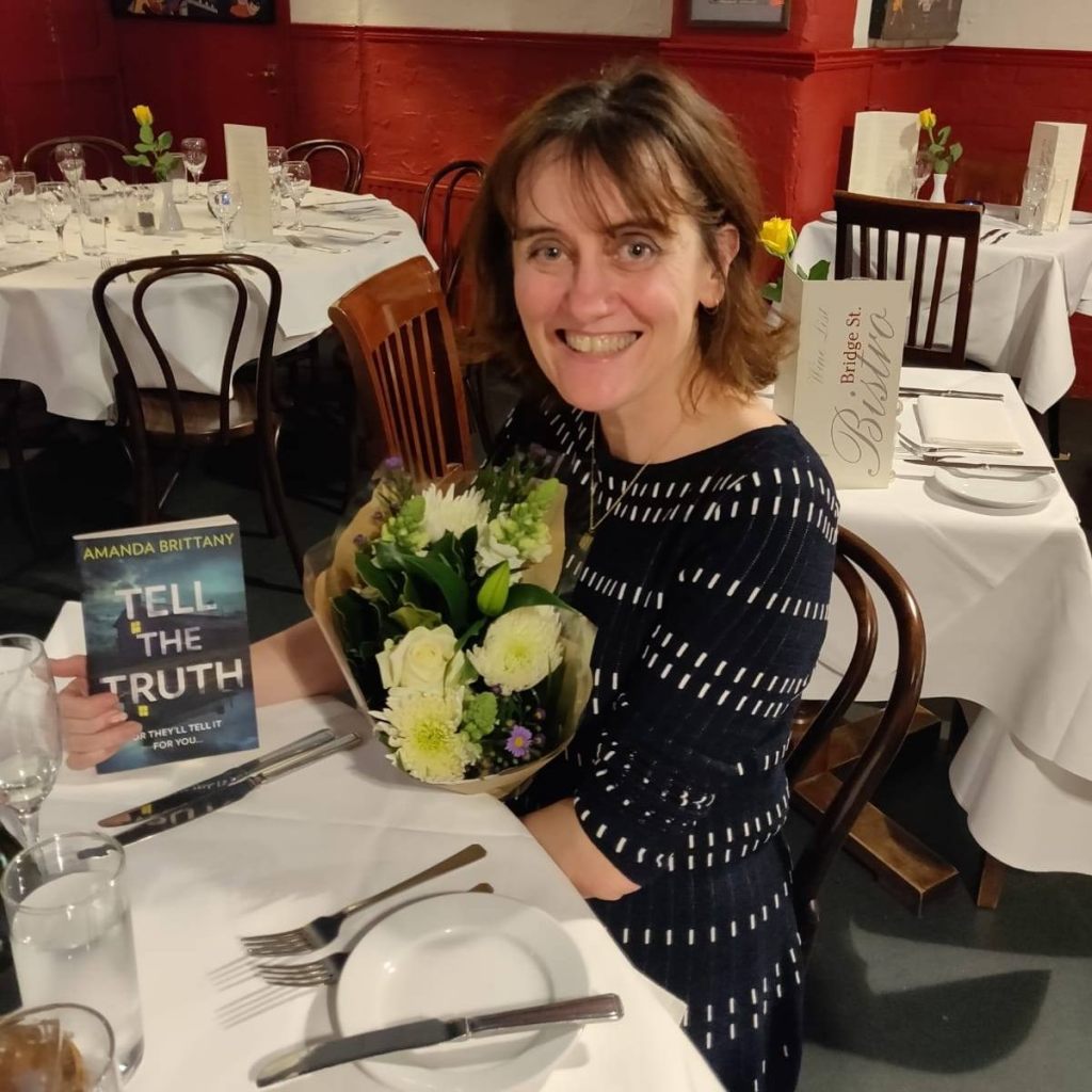 Author Amanda Brittany - Smiling middle aged woman with shoulder length brown hair wearing a black dress and seated at a table in a restaurant with a bunch of flowers and a copy of the book