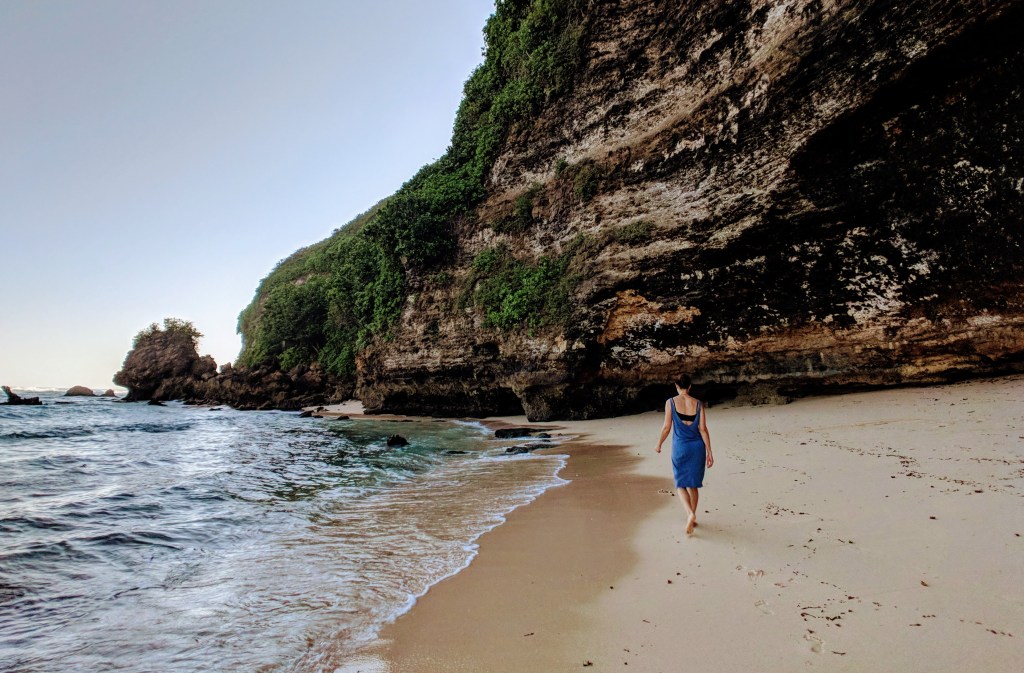 A woman in a blue dress with dark hair walking a beach, small waves on her left, rising craggy cliffs on her right, with greenery spilling over them, yellow sand under her feet.