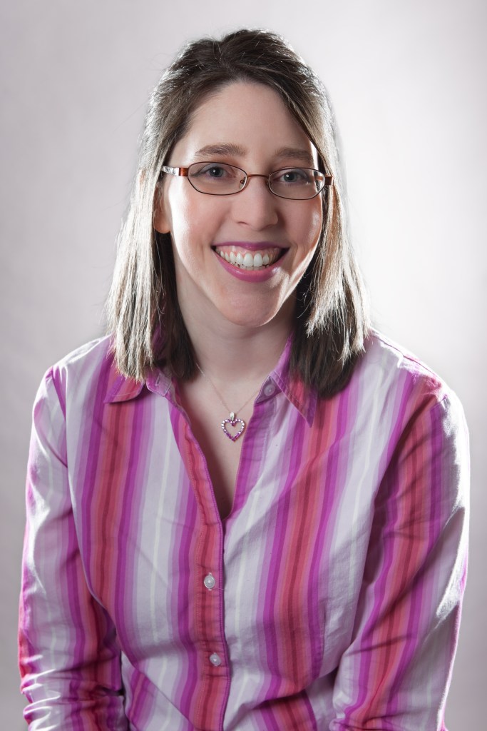 A smiling woman with straight brown hair that falls just below her shoulders. She is wearing pink lipstick, a stripy shirt (vertical, various shares of pink) and glasses.