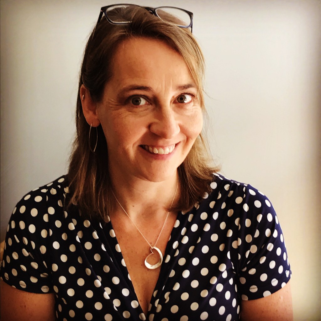 Smiling woman with shoulder length medium brown hair, held back with reading glasses on top of her head, wearing a polka-dotted black and white top and a silver heart pendant. 